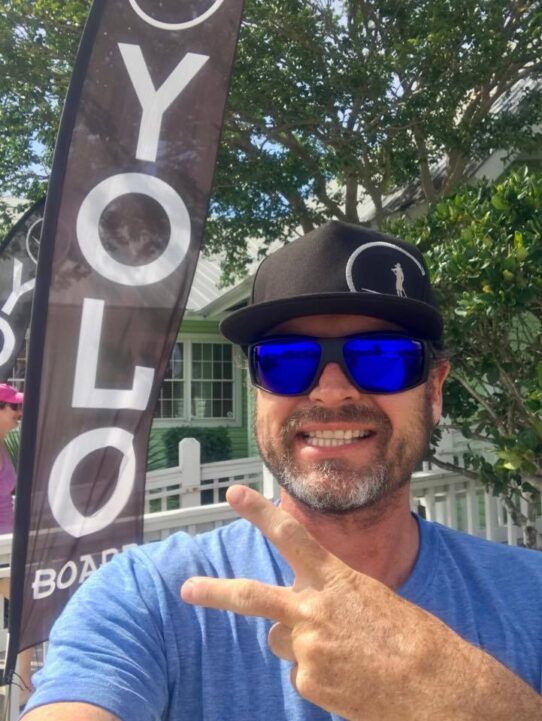 Descubre la Vida Bomber 34 Miembro del equipo de paddle board de YOLO dando un signo de paz mientras usa Bomber gafas de sol flotantes de espejo azul