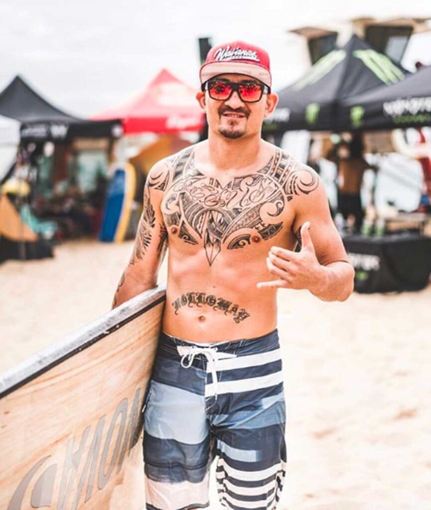 Descubre la Vida Bomber 7 Tatuado Bomber Team Surfer dando un shaka mientras sostiene su tabla en Nicaragua